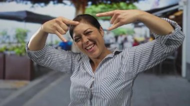 İspanyol kadın geniş bir şekilde gülümsüyor ve iki eliyle açık hava restoranının terasında kaya boynuzları hareketi yapıyor. Eğlenceli..