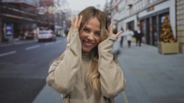 Bej kazaklı sarışın kadın, şehir caddesinde peekaboo jesti için elleriyle yüzünü kapatıyor; neşeli bir neşe.