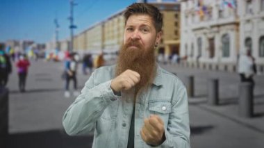 Uzun sakallı bir adam sokak ortamında orta parmağını kaldırmış, kot ceket görünür, meydan okuyor..