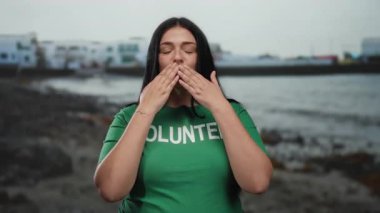 Huzurlu bir plajda gönüllü bir kadın, yeşil gönüllü gömleği giymiş, kameraya bir öpücük yolluyor sahilin manzaralı arka planına, somutlaştırıcı ilgiye ve toplum ruhuna karşı..