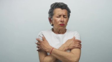 Gri saçlı yaşlı bir kadın, beyaz bir gömlek giyip beyaz bir arka plana yaslanmış, huzursuzluk ve soğukluğu ifade eden, soğuktan titriyor..