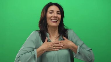 Yeşil bluzlu kadın kolunu uzatırken gülümser ve elini göğsüne parlak yeşil bir stüdyo duvarına koyar..