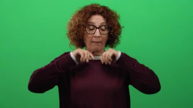 Yeşil duvara yaslanmış kıvırcık saçlı, yeşil arka planda gözlüklü ve mor kazaklı bir kadın..