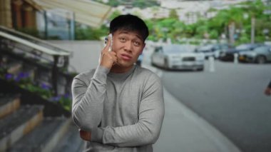 Gri kazaklı genç adam şehir caddesinde akıllı telefonla konuşuyor. Etrafı arabalar ve yeşilliklerle çevrili..
