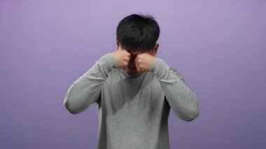 Gri kazaklı genç adam, yorgun ya da stresli görünen mor arka plana gözlerini sürterek, minimalist bir ortamda yorgunluk ya da endişeye işaret ediyor..