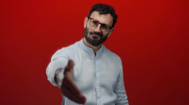 Sakallı ve gözlüklü bir adam canlı kırmızı arka plana karşı el sıkışmak için uzatıyor selamlamayı simgeliyor ve olumlu bir mesleki bağlamda hoş geldiniz..