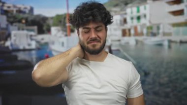 Sakallı genç bir adam, arka planda tekneler ve gemilerle resim gibi bir limanda boyun rahatsızlığı yaşıyor. Muhtemelen stresi ya da dışarıdaki acıyı tasvir ediyor..