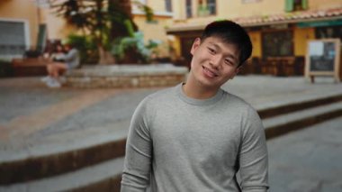 Şehrin arka planında neşeli bir şekilde gülümseyen genç bir adam, şehir manzarasını günlük atmosferiyle, güneşli günüyle ve rahat ifadesiyle yakalıyor..
