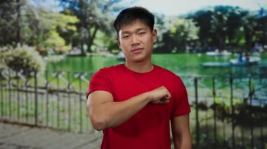 Kırmızı gömlekli genç adam yeşil ağaçlar ve dingin göl arka planıyla güneşli bir park ortamında yumruğunu uzatarak açık havada dostça bir hava yaratıyor..