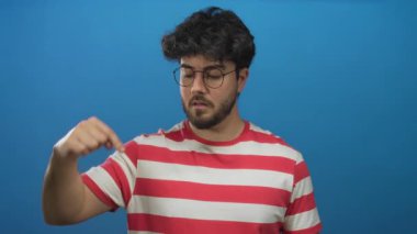Sakallı, gözlüklü ve kırmızı çizgili tişörtlü genç adam canlı mavi bir arka plana bakıyor, etkileyici jestler ve düşünceli bir yüz ifadesi sergiliyor..