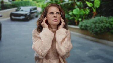 Şehir dışından bir kadın, arka plandaki şehir manzarasından rahatsız bir şekilde kulaklarını kapatıyor. Bitkiler ve araçlar, dinamik bir şehir manzarası yaratıyor..