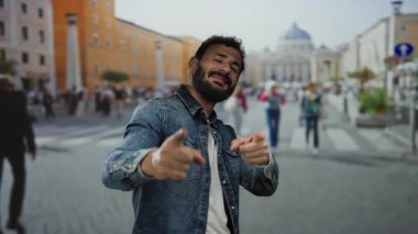 Kot ceketli İspanyol adam gülümsüyor ve arka planda bulanık yayalar ve renkli binalar olan kalabalık bir caddeyi işaret ediyor..