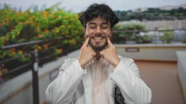 Beyaz önlüklü, sakallı genç adam güneşli bir günde açık havada gülümsüyor. Parlak yeşillik ve açık gökyüzü pozitif bir hava izlenimi veriyor..