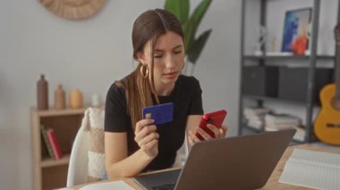 Binadaki dizüstü bilgisayarın yanındaki ahşap masaya yaslanırken kadının elinde kredi kartı ve akıllı telefon var; alışveriş heyecanı.