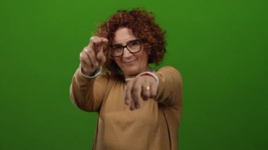 Yeşil arka planda barış jesti yapan, orta yaşlı, kıvırcık saçlı, gözlüklü ve süveterli bir kadın, canlı duvara karşı izole edilmiş, pozitifliğini ve güvenini ifade ediyor..