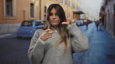 Şehirdeki genç İspanyol kadın, elinde kamerayla zavallı jestler yapıyor, açık hava ortamında gri bir kazak giyiyor ve arka planda bulanıklık var..