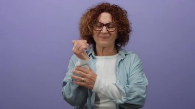 Mor duvarın önünde bileği ağrıyan bir kadın gözlük takıyor ve mavi bir gömlek giyiyor. Yüzünde endişeli bir ifade var. İçeriden izole edilmiş..