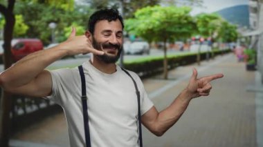 Sakallı İspanyol bir adam, şehir sokağında gülerken gündüz vakti arka planda ağaçlarla arama hareketi yapıyor..