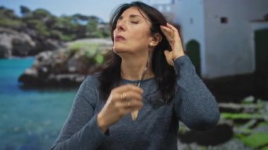 Orta yaşlı İspanyol kadın, beyaz binanın önündeki gök mavisi denizin yanındaki kayalık sahilde alnına dokunuyor. Stres..