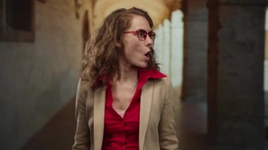 Tarihi bir üniversite koridorunda el kol hareketi yapan, şaşırdığını ve merakını dile getiren, kırmızı gözlük takan ve kapalı bir akademik ortamda bej ceket giyen bir kadın..