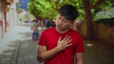 Kırmızı tişörtlü genç adam şehir caddesinde duruyor. Duygularını ifade ediyor. Göğsünde bulanık insanlar ve açık hava ışıkları altında ağaçlar var.