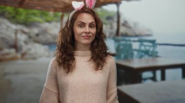 Tavşan kulaklı bir kadın, güneşli bir günde açık mavi gökyüzünün altında, kayalık kıyı şeridi ve sakin deniz tarafından çerçevelenmiş, deniz kenarındaki kafede masa ve sandalyelerle gülümsüyor..