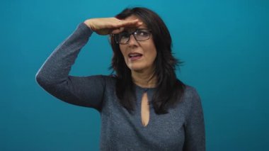 Gözlük takan bir kadın mavi stüdyoda elleri ile gözleri gölgeliyor..
