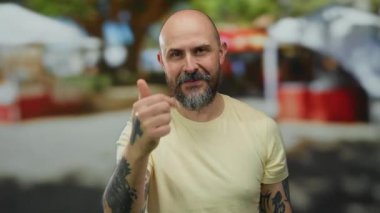 Güneşli bir günde sokak ortamında gülümseyen bir adam sarı bir gömlek giyerken sakalını, kelliğini ve dövmelerini sergiliyor..