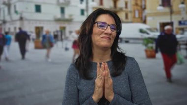 Güneşli şehir caddesinde ellerini sımsıkı bağlayıp dua eden bir kadın duruyor ve nazikçe gülümsüyor:.
