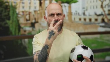 Roma harabelerinde sakallı bir adam futbol topunu tutuyor ve görünür dövmeleri olan, ciddi görünen ve dış mekâna odaklanmış bir adam..