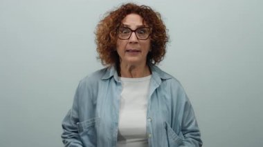 İzole edilmiş beyaz bir arka plan duvarının önünde duygularını ifade eden kadın, gözlüklü, orta yaşlı esmer bir kadını gösteriyor..