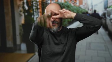 Sakallı kel bir adam barış jesti yapıyor ve arka planda bulanık insanlar ve binalar olan bir şehir sokağındaki kameraya bakıyor..