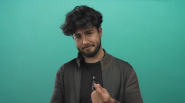 Sakallı ve üzgün yüzlü genç İspanyol bir adam canlı yeşil bir arka plana karşı poz veriyor, günlük kıyafetler ve kolye giyiyor, duyguları ve tepkileri vurguluyor..