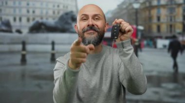 Bearded man with tattoo shows car keys on urban street backdrop suggesting transportation acquisition in vibrant city setting and cultural lifestyle.