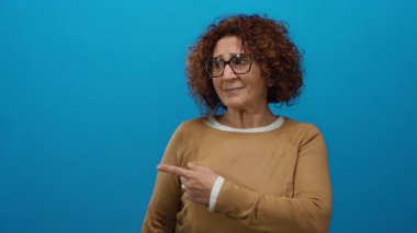 Orta yaşlı, kıvırcık saçlı, gözlüklü, parlak mavi arka planı olan, kahverengi bir süveter giyen orta yaşlı bir kadın..