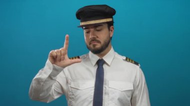Pilot üniformalı İspanyol bir adam mavi arka planda kameraya doğru bir hareket yapıyor, oyuncu bir ifadeyle mizah ve otorite sergiliyor..