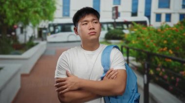 Sırt çantalı, şehir binaları ve yeşilliklerle çevrili bir sokakta duran genç adam, açık hava ortamında bir öğrenciyi işaret ediyor..