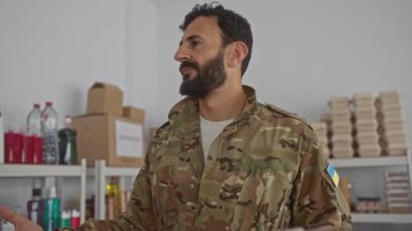 Askerî üniformalı İspanyol sakallı bir adam elinde pano, etrafı malzemeler ve Ukrayna bayrak bandıyla çevrili bir odada gönüllü çalışmalarını temsil ediyor..