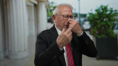 Sokaktaki kıdemli adam kötü kokuya karşı burun çimdikliyor, şehir sütunları ve yeşillikler tarafından çerçeveleniyor..