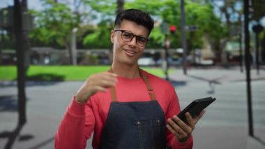 Önlüklü İspanyol bir adam şehir caddesinde tablet kullanıyor. Onay veriyor. Dışarısı canlı bir manzarayla çevrili..