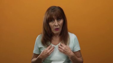 Beyaz gömlekli son sınıf öğrencisi kadın canlı turuncu arka plana şaşırmış görünüyor, dışlanmış bir ortamda duygularını ve olgun yüz hatlarını vurguluyor..