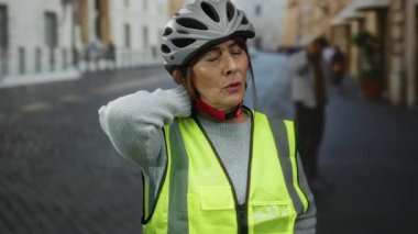 Üst düzey İspanyol kadın, kask ve güvenlik yeleği giyip kalabalık bir şehir caddesinde dikilirken boynunu ovalıyor şehir yaşamını ve kişisel güvenliğini gösteriyor..