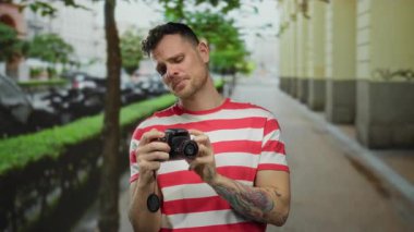 Dövmeli genç adam şehir sokağında dururken kamerayı inceliyor.