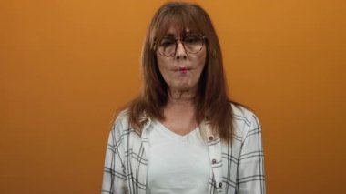 Turuncu arka planda gözlük takan, ekose gömlek giyen ve neşeli bir tavır sergileyen bir kadın..