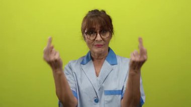 Yaşlı İspanyol kadın, gözlüklü ve üniformalı, kapalı bir yerde kararlı bir ifadeyle canlı sarı bir arkaplan karşısında meydan okuyor.