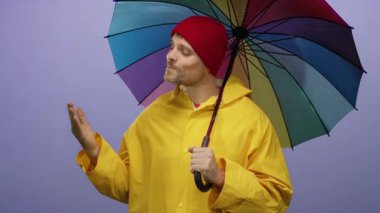 Sarı yağmurluklu genç adam mor arka plana karşı renkli bir şemsiye tutuyor. Kendine güvenen, yakışıklı bir ifadeye ve alımlı bir tavırla avuç içini açıyor..