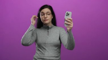 Gözlük ve gri kazak giyerken akıllı telefonlu selfie çeken bir kadın, sosyal medya aracılığıyla modern teknoloji ve kişisel ifade sergiliyor..