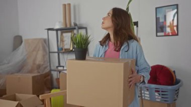 Yeni dairenin oturma odasında taşınan erzak ve dekorlarla çevrili bir kutu taşıyan kadın, genç yetişkinliği ve ev sahipliği geçişini yansıtıyor..
