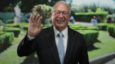 Yeşil bir park ortamında gülen ve el sallayan takım elbiseli son sınıf öğrencisi..