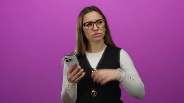 Gözlüklü genç kadın pembe arka planda akıllı telefonu tutuyor, düşünceli görünüyor ve cihazı yakından düşünüyor..